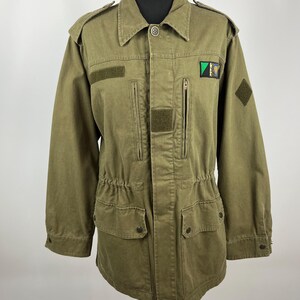 May include: Olive green military-style jacket with a button-down front, two chest pockets, and two lower pockets. Features a small patch on the upper right chest and a diamond-shaped patch on the right sleeve. The jacket has a drawstring waist and epaulettes.