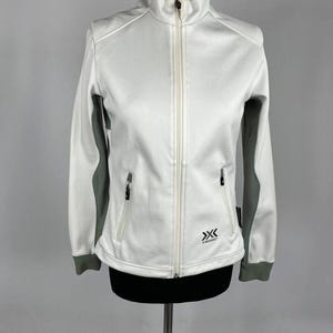 Puede incluir: Chaqueta deportiva blanca y gris con cuello alto y cremallera completa. La chaqueta tiene mangas y ribetes grises, dos bolsillos con cremallera y un pequeño logotipo en la parte delantera. La chaqueta se exhibe en un maniquí.