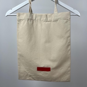 May include: A rectangular beige canvas tote bag with two matching fabric handles. The bag features a small, solid red rectangular patch centred near the bottom edge. The tote is suspended from a white plastic hanger against a neutral background.