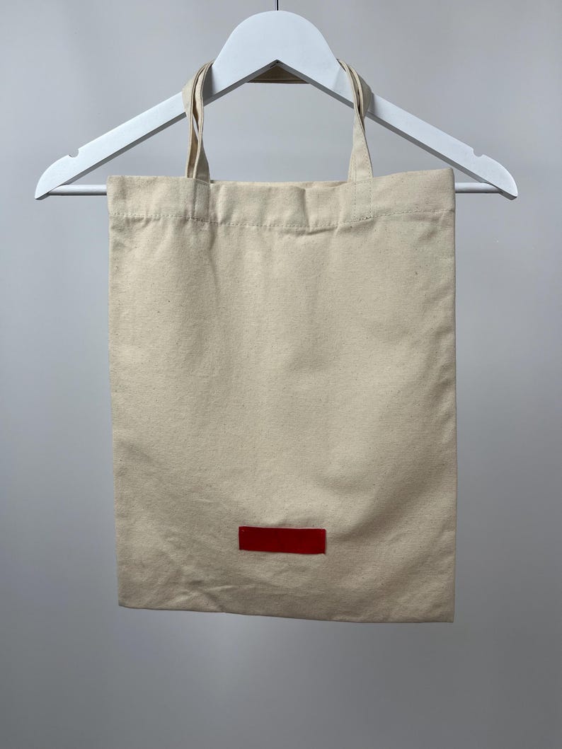 May include: A rectangular beige canvas tote bag with two matching fabric handles. The bag features a small, solid red rectangular patch centred near the bottom edge. The tote is suspended from a white plastic hanger against a neutral background.
