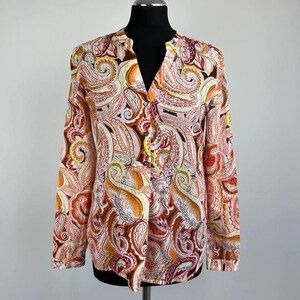 Könnte beinhalten: Eine langärmlige Bluse mit Stehkragen und V-Ausschnitt. Die Bluse hat ein farbenfrohes Paisley-Muster in Orange-, Rosa-, Gelb- und Brauntönen. Das Kleidungsstück wird auf einer schwarzen Schaufensterpuppe präsentiert.