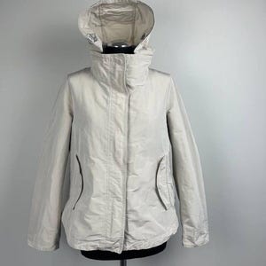 May include: A light beige hooded jacket with a high collar and two front pockets. The jacket is displayed on a mannequin against a plain background. The hood is adjustable and the jacket has a zipper closure.