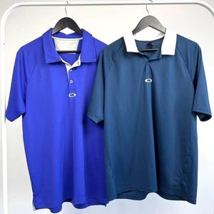 May include: Two short-sleeved polo shirts hanging on a white rack. One is a vibrant blue, the other is a deep navy with a white collar. Both shirts have a three-button placket and a small logo near the chest.