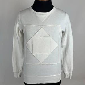 May include: A white sweatshirt with a geometric design. The long-sleeved top features a diamond-shaped patch on the front, with a smaller triangle detail at the neckline. The sweatshirt is displayed on a mannequin.