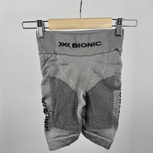 Puede incluir: Pantalones cortos deportivos grises con el texto "X BIONIC" impreso en la cintura y la pierna. Los pantalones cortos están colgados de una percha de madera. Los pantalones cortos tienen un diseño texturizado en la parte delantera y trasera.