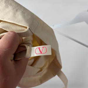 May include: A cream-coloured canvas tote bag featuring a small, rectangular fabric tag sewn into the interior seam. The tag is white with a bright pink V logo enclosed in a circle, representing the Valentino brand identity on the accessory.