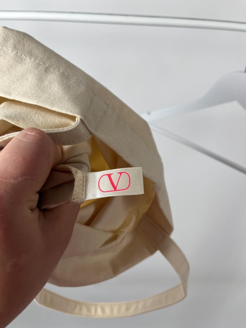 May include: A cream-coloured canvas tote bag featuring a small, rectangular fabric tag sewn into the interior seam. The tag is white with a bright pink V logo enclosed in a circle, representing the Valentino brand identity on the accessory.