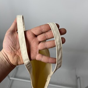 May include: A plain, off-white canvas tote bag featuring two sturdy, stitched fabric handles. The bag is constructed from a light-coloured, woven material with visible seams along the edges of the handles, providing a simple and functional design.
