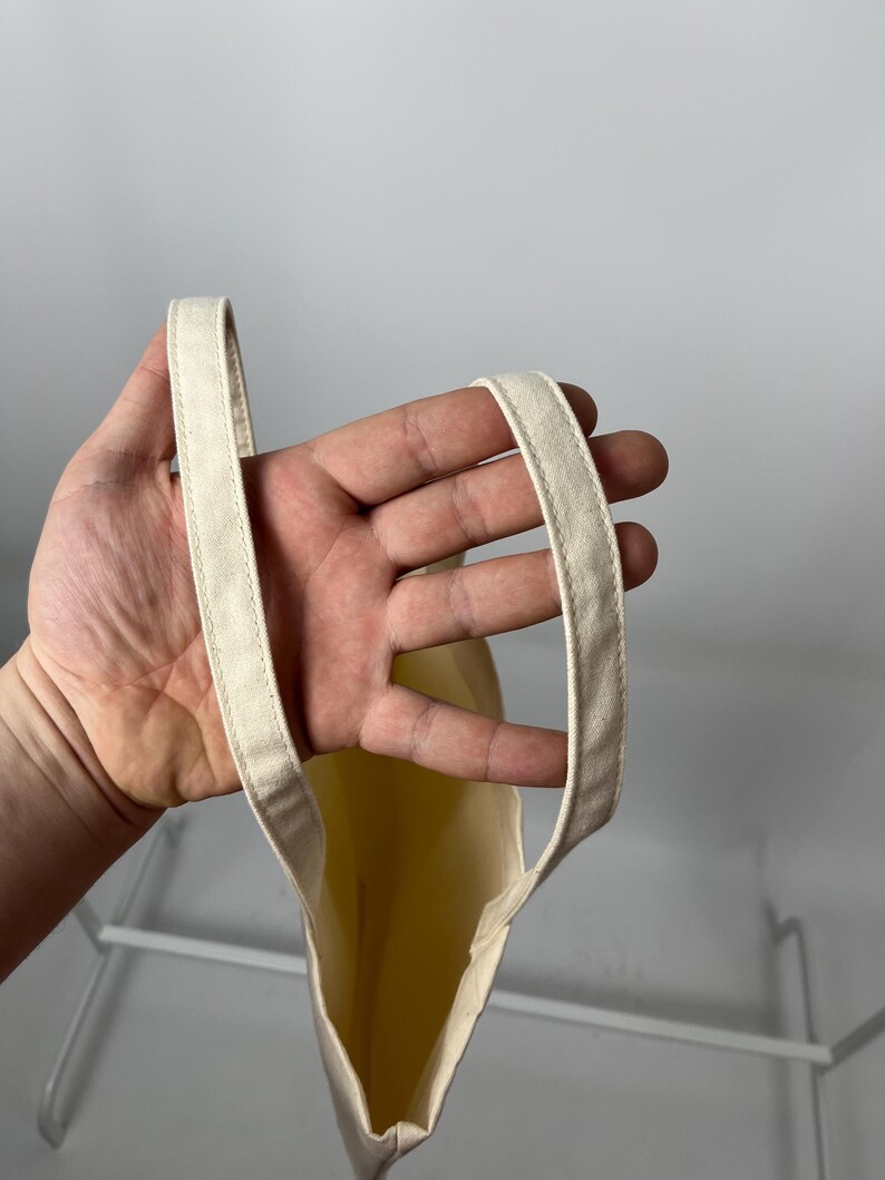 May include: A plain, off-white canvas tote bag featuring two sturdy, stitched fabric handles. The bag is constructed from a light-coloured, woven material with visible seams along the edges of the handles, providing a simple and functional design.