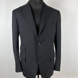 May include: A black blazer with a two-button closure, a notched lapel, and two front pockets. The blazer is displayed on a mannequin with a wooden head. The fabric appears to be a solid, dark color.
