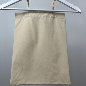 May include: A plain, rectangular, off-white canvas tote bag with two matching fabric handles. The bag is constructed from a woven textile material and is suspended from a white plastic hanger, displaying a simple, functional design.