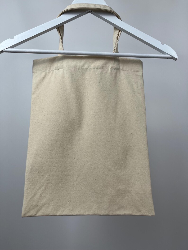 May include: A plain, rectangular, off-white canvas tote bag with two matching fabric handles. The bag is constructed from a woven textile material and is suspended from a white plastic hanger, displaying a simple, functional design.