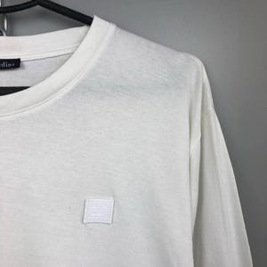 Könnte beinhalten: Weißes Langarm-T-Shirt mit Rundhalsausschnitt. Ein kleines, quadratisches, weißes Patch mit minimalistischem Gesichtsdesign ist auf die Brust genäht. Das Shirt hängt an einem schwarzen Kleiderbügel vor grauem Hintergrund.