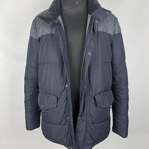 Puede incluir: Chaqueta acolchada azul marino oscuro con cuello y paneles en los hombros grises. La chaqueta tiene cremallera, dos bolsillos delanteros y un diseño acolchado. La chaqueta se muestra en un maniquí.