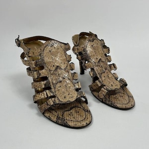 Op de afbeelding: Een paar sandalen met hoge hakken met een slangenprint in bruin- en beigetinten. De sandalen hebben meerdere bandjes en een gespsluiting. De hakken zijn ongeveer 7.6 cm hoog.