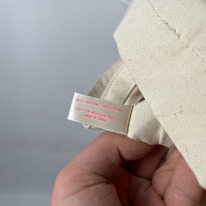 May include: A cream-coloured cotton fabric item featuring a white tag with red text that reads: BODY MATERIAL 100% COTTON, NOT FOR INDIVIDUAL SALE, MADE IN CHINA. The fabric has a visible woven texture and is folded with stitched seams.