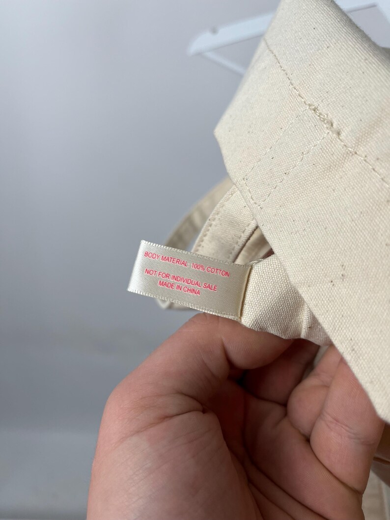May include: A cream-coloured cotton fabric item featuring a white tag with red text that reads: BODY MATERIAL 100% COTTON, NOT FOR INDIVIDUAL SALE, MADE IN CHINA. The fabric has a visible woven texture and is folded with stitched seams.