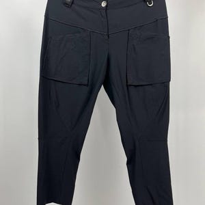 Puede incluir: Pantalones negros cortos con grandes bolsillos delanteros y cierre de botón. Los pantalones tienen cintura alta y cuelgan de una percha negra. La tela parece ser un material ligero.