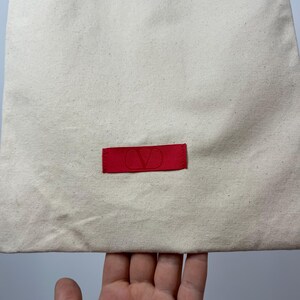 May include: A rectangular off-white canvas drawstring bag featuring a centred horizontal red fabric label. The label displays a debossed V logo in the centre. The fabric has a visible woven texture with small dark flecks throughout the material.