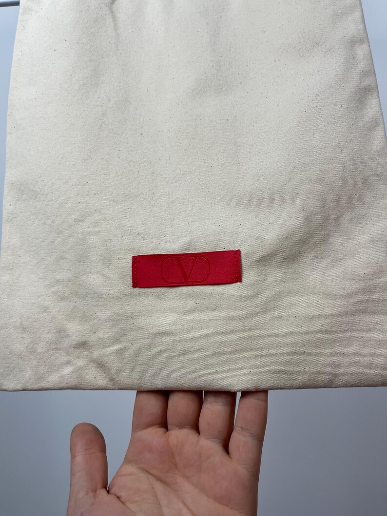 May include: A rectangular off-white canvas drawstring bag featuring a centred horizontal red fabric label. The label displays a debossed V logo in the centre. The fabric has a visible woven texture with small dark flecks throughout the material.