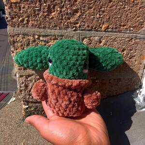 May include: A green and brown plush toy shaped like a small potted plant. The toy has a round head with two small ears and a brown pot.