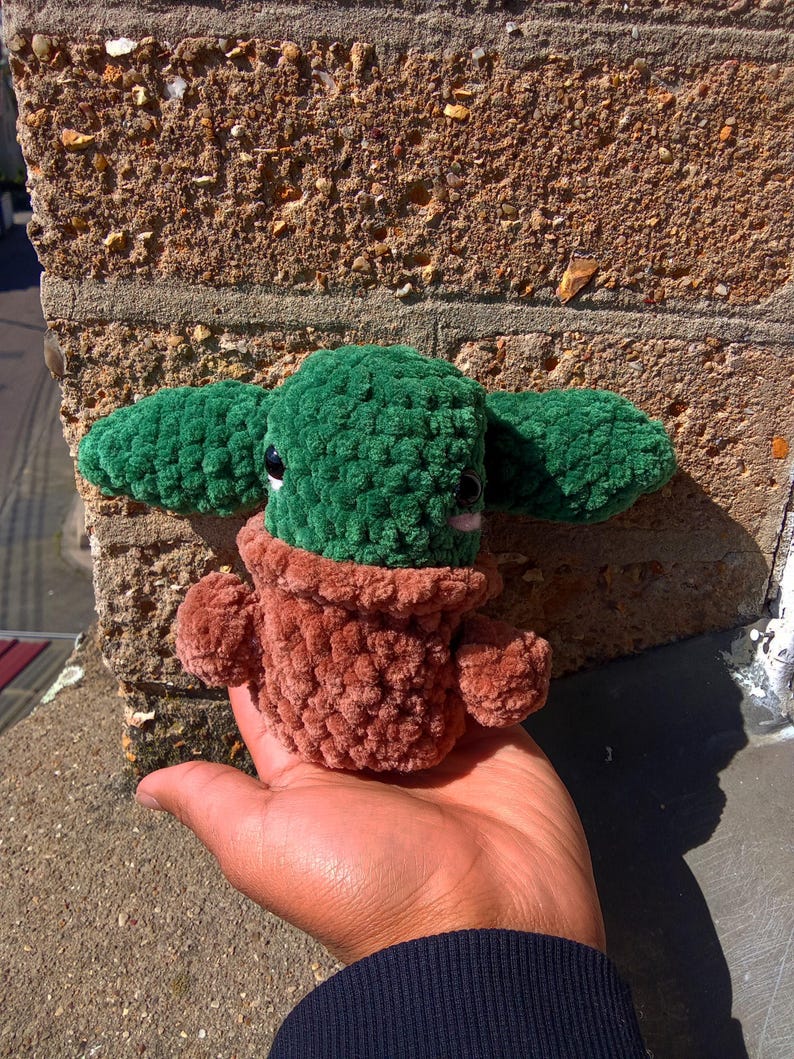 May include: A green and brown plush toy shaped like a small potted plant. The toy has a round head with two small ears and a brown pot.