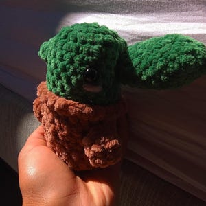 May include: A green and brown crocheted plush toy resembling a character from the Star Wars franchise. The toy is being held in a person's hand.