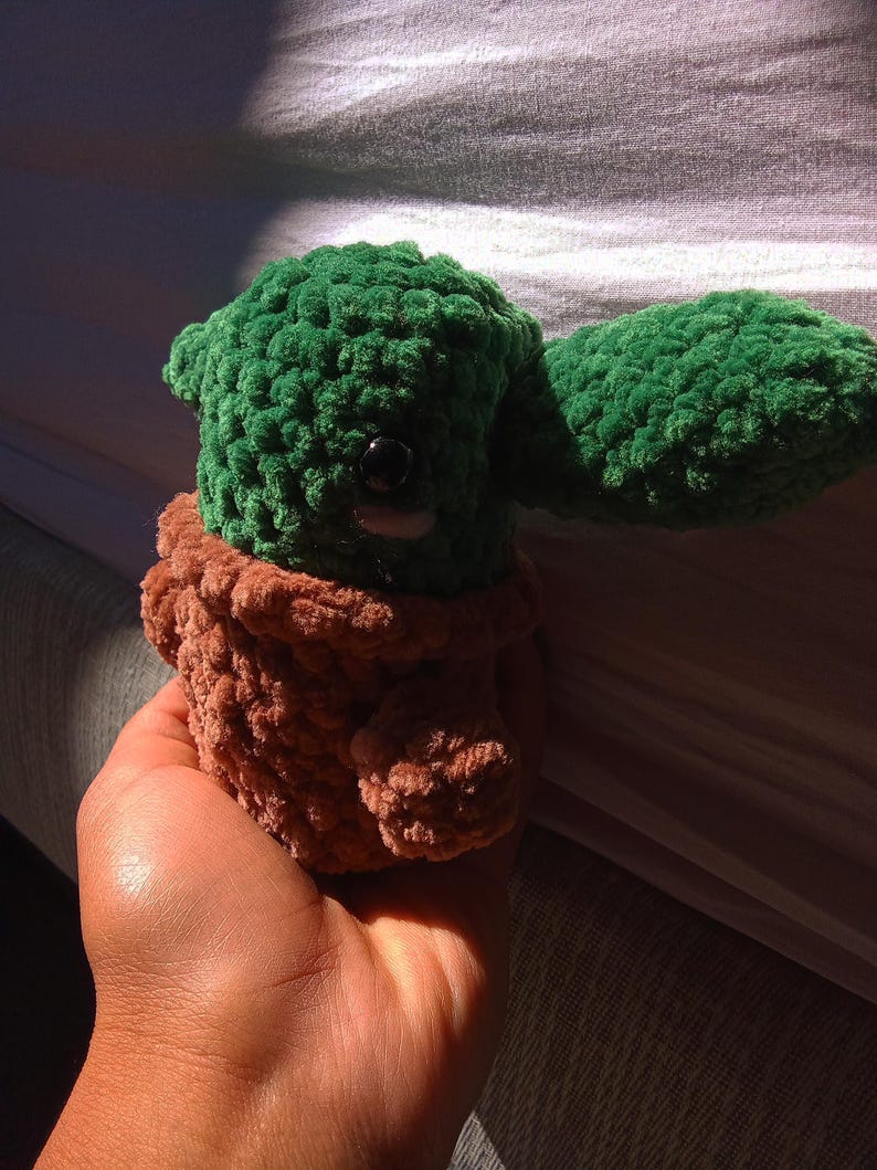 May include: A green and brown crocheted plush toy resembling a character from the Star Wars franchise. The toy is being held in a person's hand.