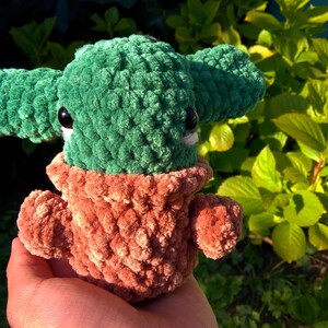 May include: A green and brown crocheted plush toy in the shape of a character with large eyes. The toy is being held in a person's hand.