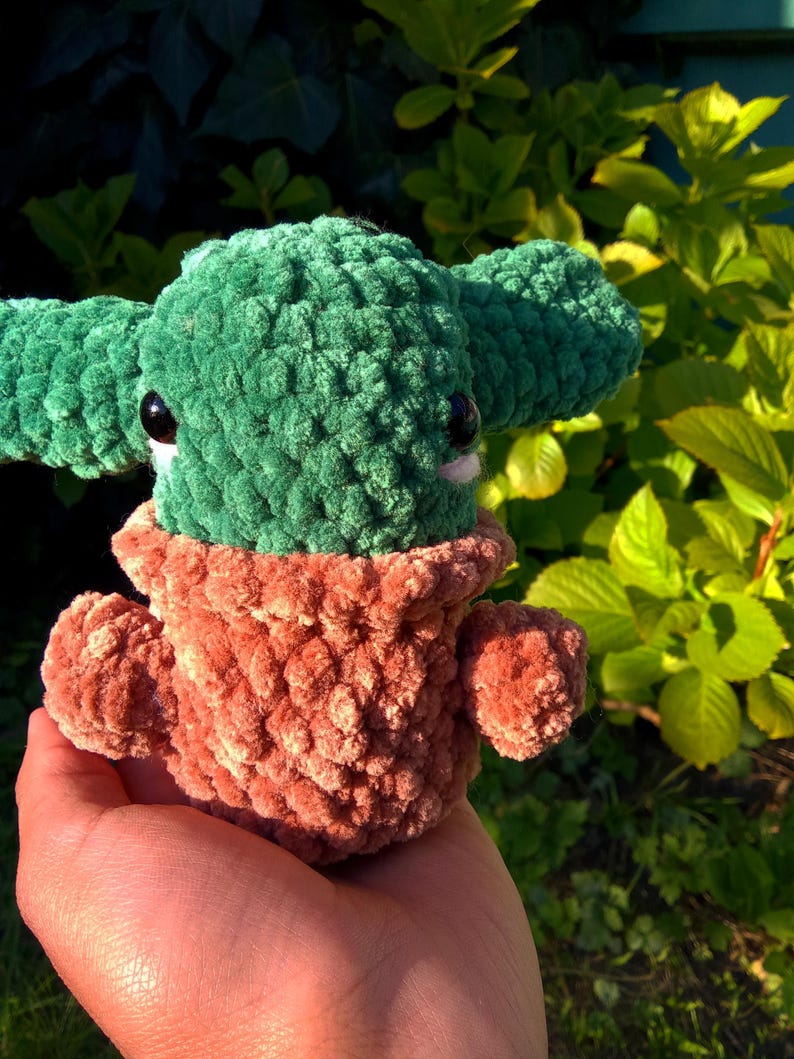 May include: A green and brown crocheted plush toy in the shape of a character with large eyes. The toy is being held in a person's hand.