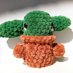 May include: A green crocheted plush toy shaped like a potted plant with large eyes and a brown crocheted pot.