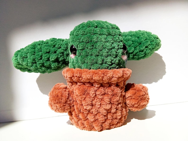 May include: A green crocheted plush toy shaped like a potted plant with large eyes and a brown crocheted pot.