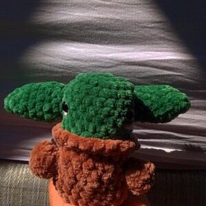 May include: A green and brown crocheted plush toy resembling a character from the Star Wars franchise. The toy has large ears, a small mouth, and big eyes.