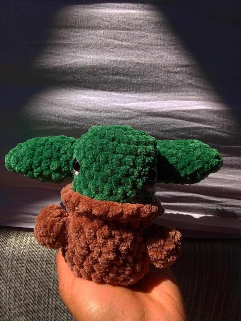 May include: A green and brown crocheted plush toy resembling a character from the Star Wars franchise. The toy has large ears, a small mouth, and big eyes.