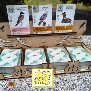 May include: A wooden card holder with a bird design, holding three bird cards and four stacks of cards. The bird cards have illustrations of different birds and text. A small yellow square with puzzle pieces is in the corner.