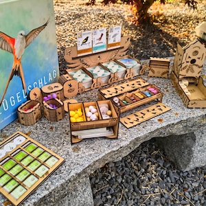 Può includere: Un set di giochi da tavolo in legno con vari componenti, tra cui una scatola con il titolo "FlügelSchlag" e un'illustrazione di un uccello. Il set include vassoi, contenitori e pezzi del gioco in legno, il tutto disposto su una superficie di pietra.