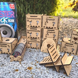 K&ouml;nnte beinhalten: Eine Ansammlung von Holzkisten und Spielkomponenten. Die Kisten sind mit verschiedenen Themen wie "Rock", "Summerparty" und "Movies & TV" gelasert. Eine Spielbox mit dem Titel "Hitster Rock Bob!" ist im Hintergrund zu sehen.