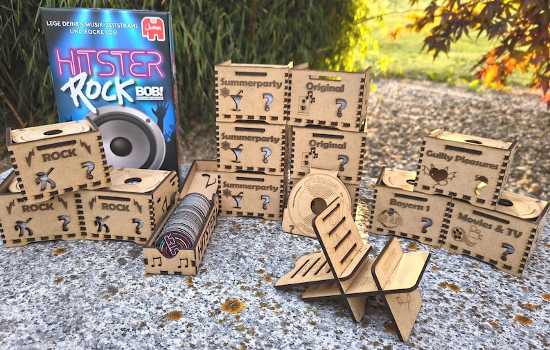 K&ouml;nnte beinhalten: Eine Ansammlung von Holzkisten und Spielkomponenten. Die Kisten sind mit verschiedenen Themen wie "Rock", "Summerparty" und "Movies & TV" gelasert. Eine Spielbox mit dem Titel "Hitster Rock Bob!" ist im Hintergrund zu sehen.