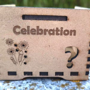 K&ouml;nnte beinhalten: Kleine, hellbraune Holzkiste mit dem Wort "Celebration" auf der Vorderseite eingraviert. Die Kiste hat ein Feuerwerks- und Stern-Design, einen Fragezeichen-Ausschnitt und eine rechteckige &Ouml;ffnung oben.