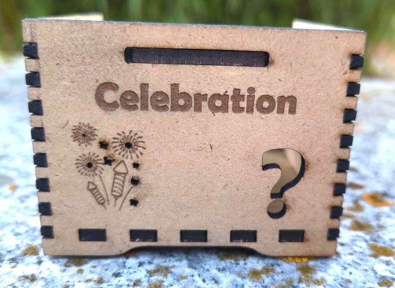 K&ouml;nnte beinhalten: Kleine, hellbraune Holzkiste mit dem Wort "Celebration" auf der Vorderseite eingraviert. Die Kiste hat ein Feuerwerks- und Stern-Design, einen Fragezeichen-Ausschnitt und eine rechteckige &Ouml;ffnung oben.