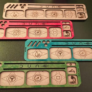 Könnte beinhalten: Vier Holzspieltabletts für das Brettspiel Dune. Jedes Tablett hat eine andere Farbe: Braun, Rot, Türkis und Grün. Die Tabletts haben Fächer für Würfel, Spielsteine und Karten. Die Tabletts sind mit "Dune" beschriftet und haben verschiedene Symbole.