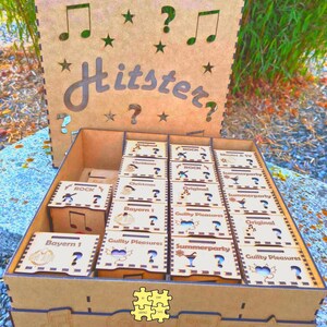 K&ouml;nnte beinhalten: Eine braune Holzspielbox mit dem ausgeschnittenen Wort "Hitster". Im Inneren befinden sich kleinere Boxen mit Aufschriften wie "Rock" und "Summerparty". Das Design umfasst Noten, Sterne und Fragezeichen, die in das Holz geschnitten sind.