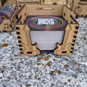 K&ouml;nnte beinhalten: Eine Holzkiste mit lasergeschnittenen Gitarrenmustern an den Seiten, die einen Stapel Karten enth&auml;lt. Die Karten haben ein farbenfrohes, kreisf&ouml;rmiges Design mit einem QR-Code. Weitere Holzkisten mit &auml;hnlichen Designs sind zu sehen.