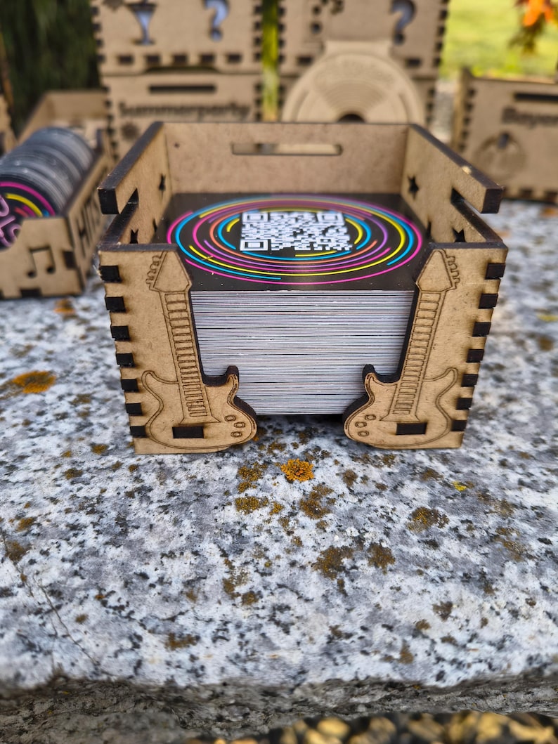 K&ouml;nnte beinhalten: Eine Holzkiste mit lasergeschnittenen Gitarrenmustern an den Seiten, die einen Stapel Karten enth&auml;lt. Die Karten haben ein farbenfrohes, kreisf&ouml;rmiges Design mit einem QR-Code. Weitere Holzkisten mit &auml;hnlichen Designs sind zu sehen.
