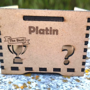 K&ouml;nnte beinhalten: Kleine, rechteckige Holzkiste mit dem Wort "Platin" auf der Vorderseite eingraviert. Die Box hat ein ausgeschnittenes Fragezeichen und eine Troph&auml;e mit dem Text "The Best!" auf der Vorderseite. Die Box hat einen Griff oben und dekorative schwarze Quadrate an den Kanten.