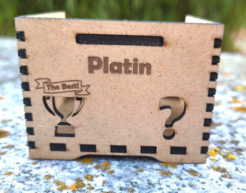 K&ouml;nnte beinhalten: Kleine, rechteckige Holzkiste mit dem Wort "Platin" auf der Vorderseite eingraviert. Die Box hat ein ausgeschnittenes Fragezeichen und eine Troph&auml;e mit dem Text "The Best!" auf der Vorderseite. Die Box hat einen Griff oben und dekorative schwarze Quadrate an den Kanten.