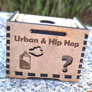 K&ouml;nnte beinhalten: Hellbraune Holzkiste mit den Worten "Urban & Hip Hop" auf der Vorderseite. Die Kiste zeigt eine Spr&uuml;hdosen-Grafik und ein Fragezeichen. Die Kiste ist quadratisch.