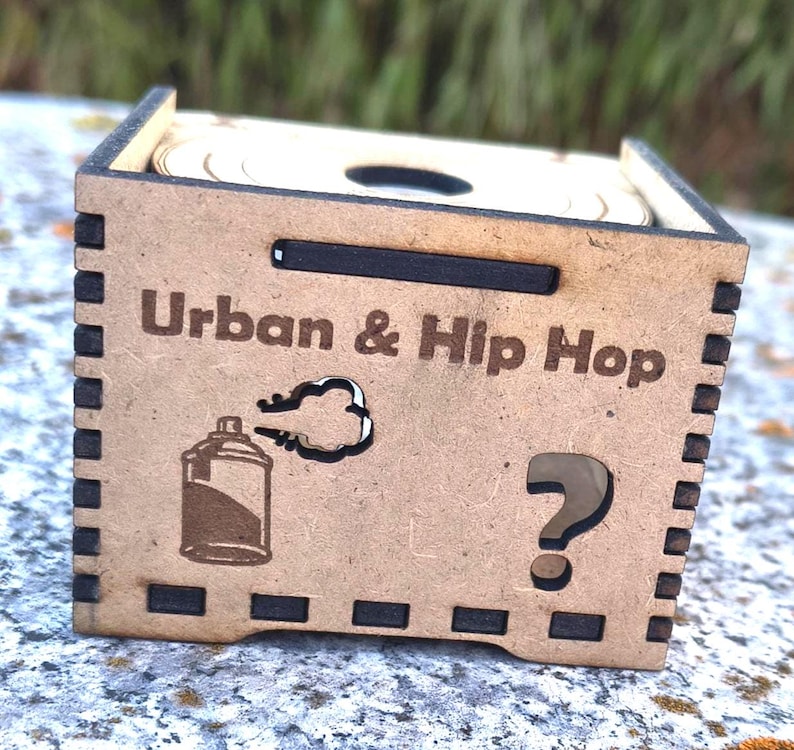 K&ouml;nnte beinhalten: Hellbraune Holzkiste mit den Worten "Urban & Hip Hop" auf der Vorderseite. Die Kiste zeigt eine Spr&uuml;hdosen-Grafik und ein Fragezeichen. Die Kiste ist quadratisch.