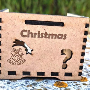 K&ouml;nnte beinhalten: Kleine, hellbraune Holzkiste mit dem Wort "Christmas" auf der Vorderseite eingraviert. Die Box zeigt einen Sternschnuppen, Glocken und ein Fragezeichen-Ausschnitt. Die Seiten haben schwarze Akzente.