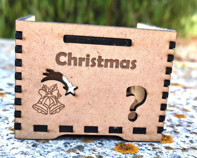 K&ouml;nnte beinhalten: Kleine, hellbraune Holzkiste mit dem Wort "Christmas" auf der Vorderseite eingraviert. Die Box zeigt einen Sternschnuppen, Glocken und ein Fragezeichen-Ausschnitt. Die Seiten haben schwarze Akzente.